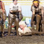 Old lady participant crawling through the mud while here teammates cheering for her